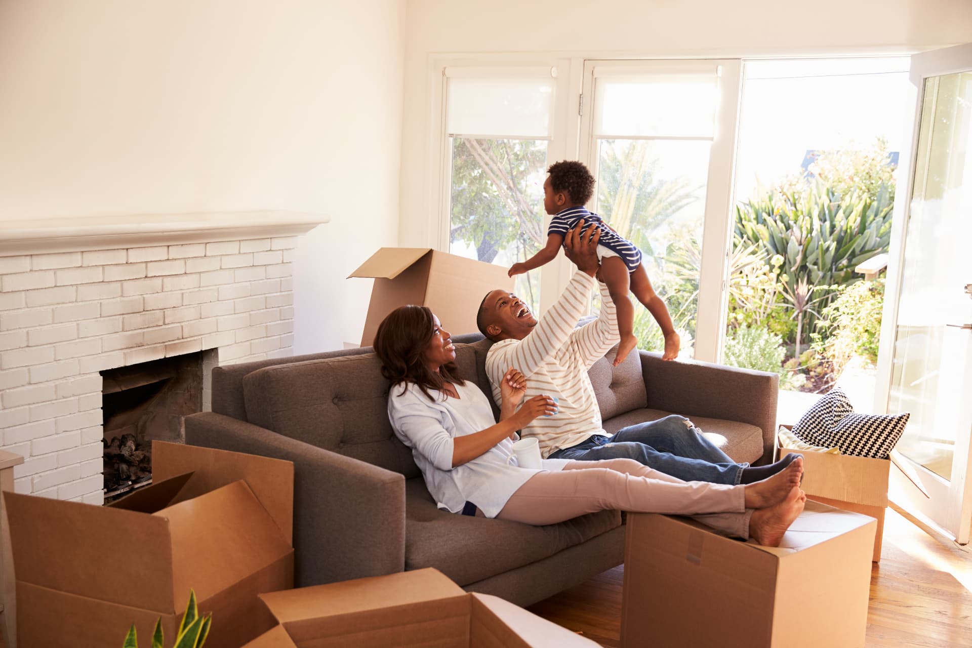 A joyful family celebrates moving into their new home, a father lifting his baby in a sunlit living room filled with moving boxes